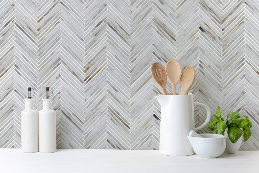 A modern kitchen countertop features two white oil and vinegar bottles, a white jug with wooden utensils, fresh basil, and a white mortar and pestle, set against a Luna Chevron Glass Tile herringbone backsplash.