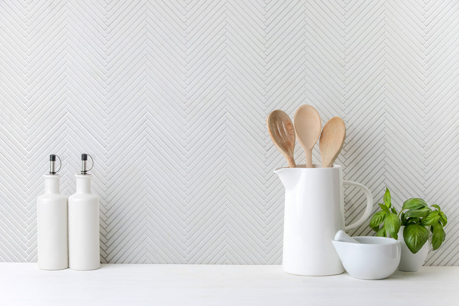 A minimal kitchen countertop features two white oil dispensers, a white pitcher with wooden spoons, a green basil plant, and a white mortar and pestle set against the Luna Chevron Glass Tile backsplash.