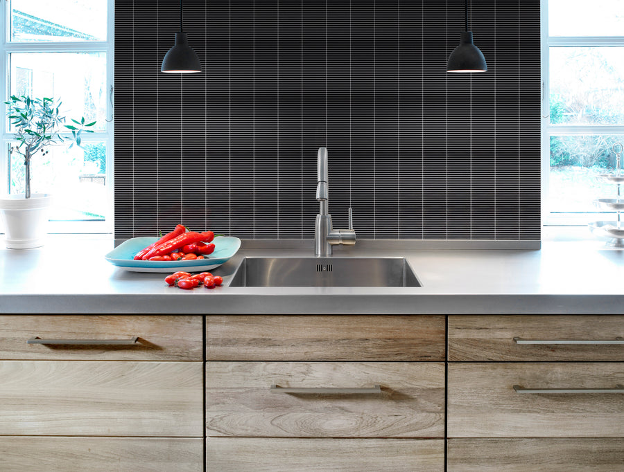 Modern kitchen with light wood cabinets and stainless steel sink features a striking Luna Kit Kat Glass Tile backsplash. Two black pendant lights hang above, while a blue plate of red bell peppers sits by the sunlit window.