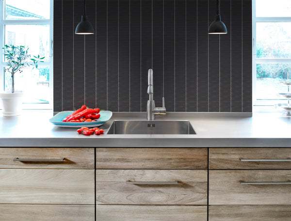 Modern kitchen with light wood cabinets and stainless steel sink features a striking Luna Kit Kat Glass Tile backsplash. Two black pendant lights hang above, while a blue plate of red bell peppers sits by the sunlit window.