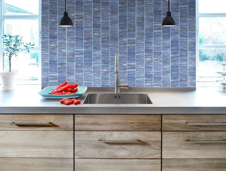 Modern kitchen with light wood cabinets, a stainless steel sink, Luna Kit Kat Glass Tile backsplash, and two black pendant lights. A turquoise tray with red bell peppers sits on the counter near a potted plant by the window.