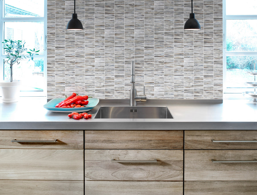 Modern kitchen with light wood drawers, stainless sink, gray countertop, and two black pendant lights. A Luna Kit Kat Glass Tile backsplash adds flair behind a blue plate with red peppers near a bright window.