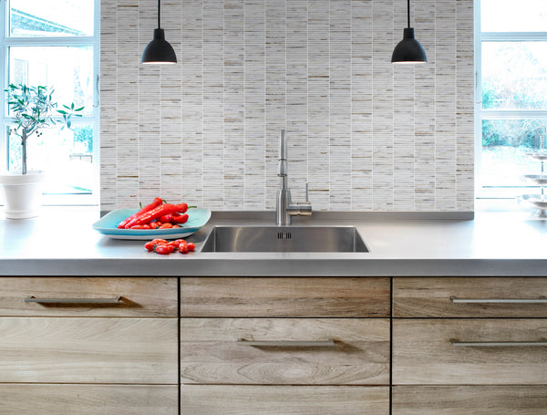 Modern kitchen with wood drawers, stainless steel sink, and two black pendants. A blue plate of red peppers sits near a potted plant by large windows, accented by Luna Kit Kat Glass Tile for added elegance.