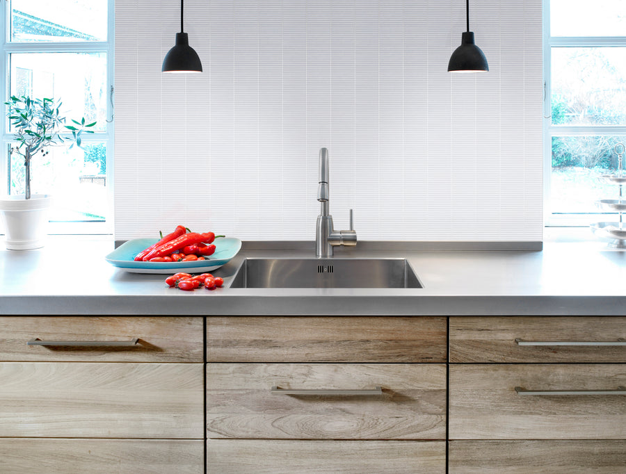 Modern kitchen with light wood cabinets, stainless steel sink, and minimalist faucet. Two black pendant lights hang above the counter, highlighting the Luna Kit Kat Glass Tile backsplash. Large windows fill the space with natural light.