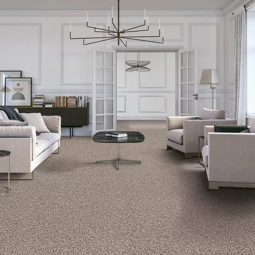 A modern living room with light-colored armchairs, a sofa, a round coffee table, and a chandelier. The room features light carpeting, white paneled walls, large windows, and double French doors opening to another space.