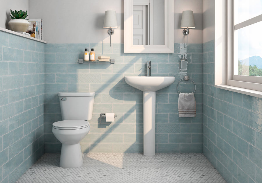A modern bathroom with blue wall tiles, a white toilet, a pedestal sink, a towel ring with a towel, wall-mounted shelves with toiletries, a mirror, and two sconces. Natural light enters through a window.