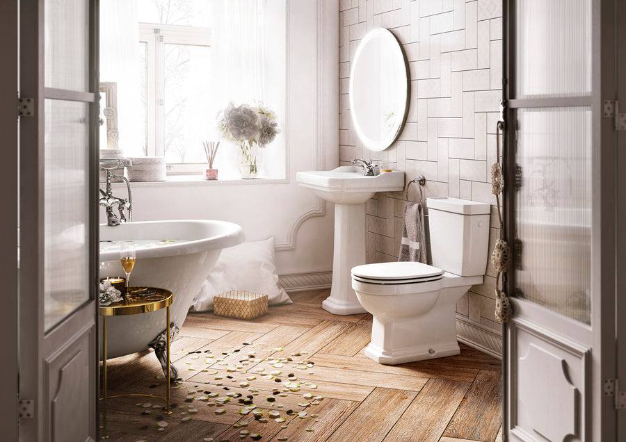 Bright, elegant bathroom with a clawfoot tub, pedestal sink, and toilet. Rose petals and a wine glass sit by the tub. Light flows through a window, highlighting flowers, candles, and a circular mirror above the sink.