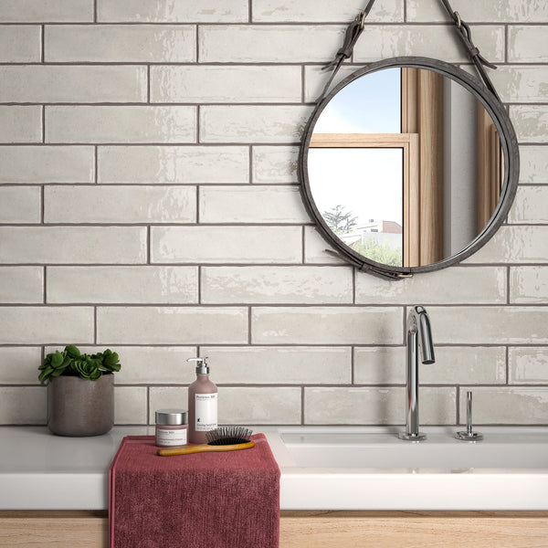 A modern bathroom with glossy subway tiles, a round mirror, a sink with a silver faucet, a small potted plant, soap dispenser, grooming items, and a folded red towel on the white counter.