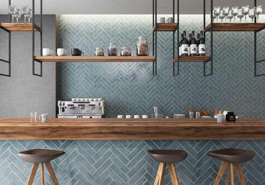 A modern coffee bar with a wood countertop, three brown stools, blue herringbone tile wall, wooden shelves holding jars, bottles, and glasses, and a silver espresso machine on the counter.