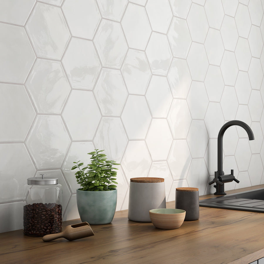 A kitchen countertop with a jar of coffee beans, a potted plant, ceramic containers, and a black faucet, set against a glossy white hexagonal tile backsplash.