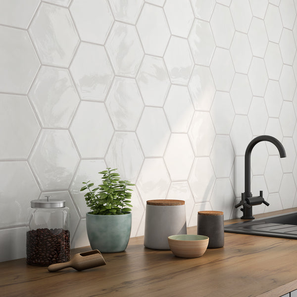 A kitchen countertop with a jar of coffee beans, a potted plant, ceramic containers, and a black faucet, set against a glossy white hexagonal tile backsplash.