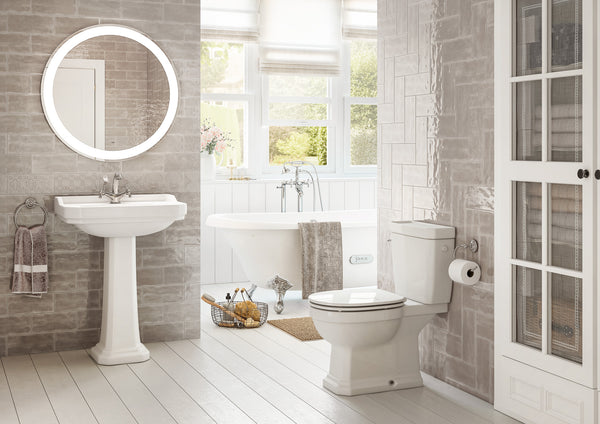 Bright, modern bathroom with a pedestal sink, round illuminated mirror, toilet, freestanding bathtub, wicker basket, towel, and large windows letting in natural light. Walls and floors are light-colored and clean.