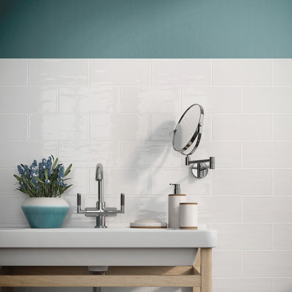 A modern bathroom sink with a chrome faucet, a small round mirror, a soap dispenser, and a light blue pot with purple flowers on a wooden vanity against a white tiled wall with a teal upper section.
