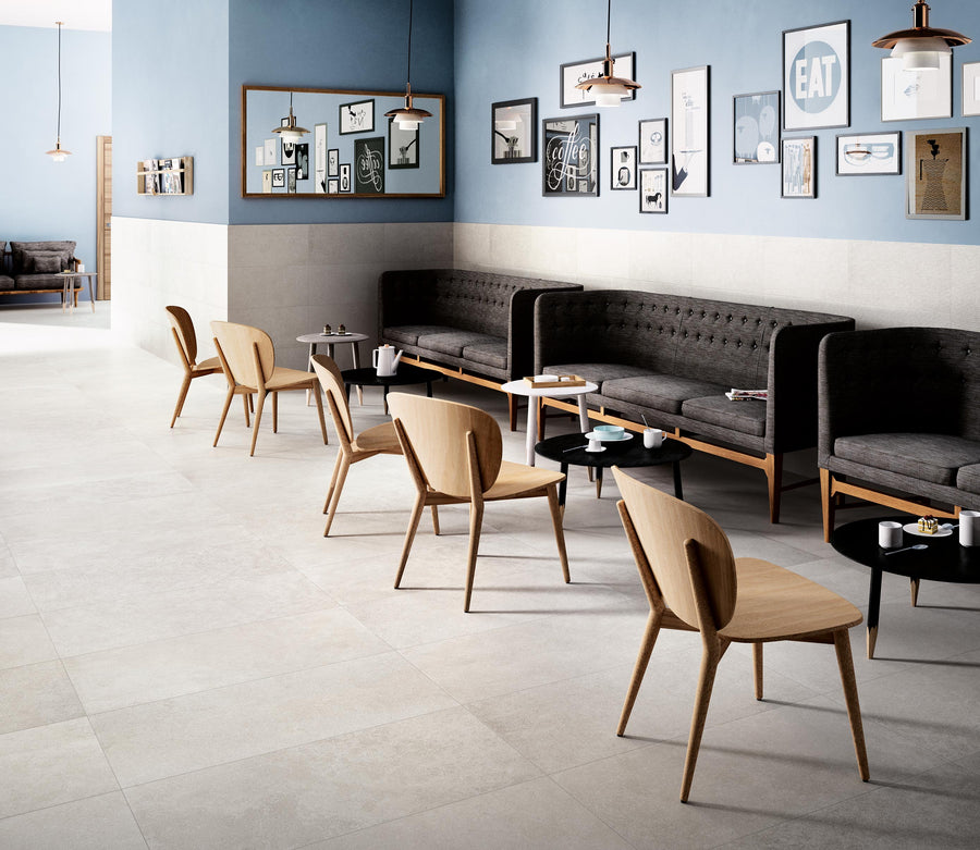 A modern cafe interior with light tiled floors, wooden chairs, black cushioned benches along blue and white walls, and framed artwork and photos hanging above the seating area. Small round tables hold cups and plates.