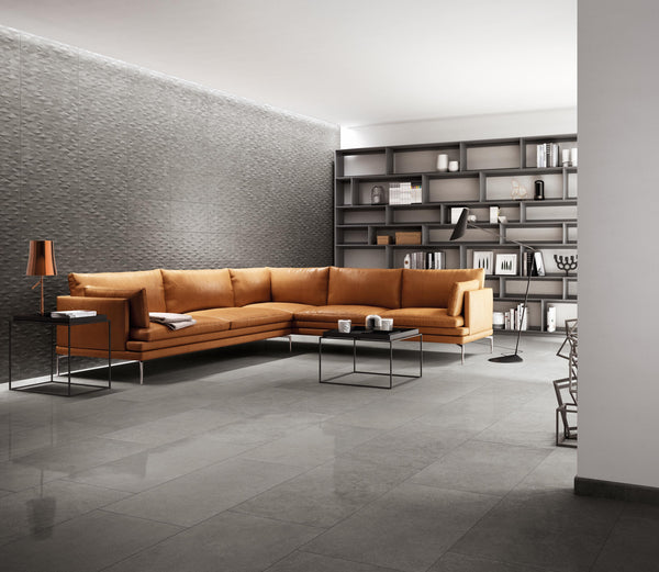 Modern living room with a large orange sectional sofa, black metal coffee tables, textured gray wall, built-in shelves with books and decor, and gray tile flooring. Warm lighting adds a cozy atmosphere.