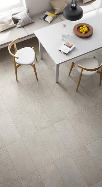 A modern dining area with light beige floor tiles, a white table with two wooden chairs, a bowl of lemons, and cushioned seating by a window. Natural light brightens the minimalist space.