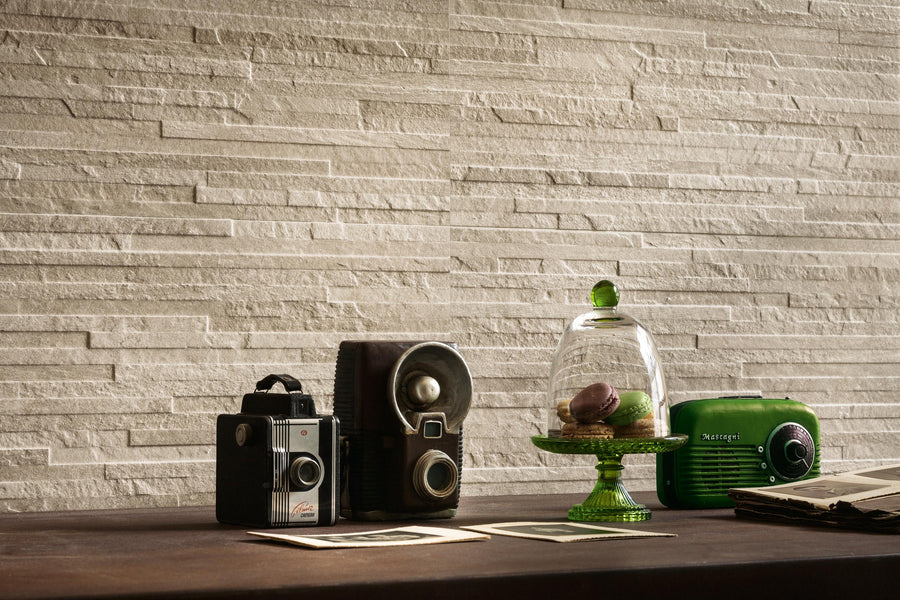 A table with two vintage cameras, old photos, a green glass stand with macarons under a dome, and a green retro radio, set against a textured beige stone wall.