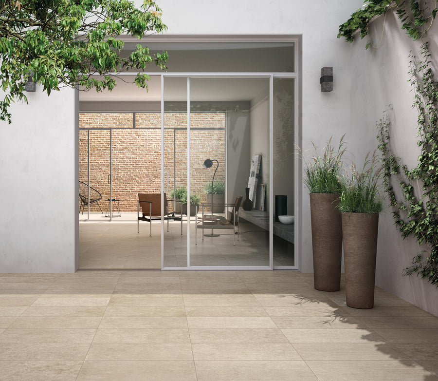 A modern patio with large beige tiles, sliding glass doors, tall planters with green plants, and a view into a stylish indoor living area with natural light and minimalistic decor.