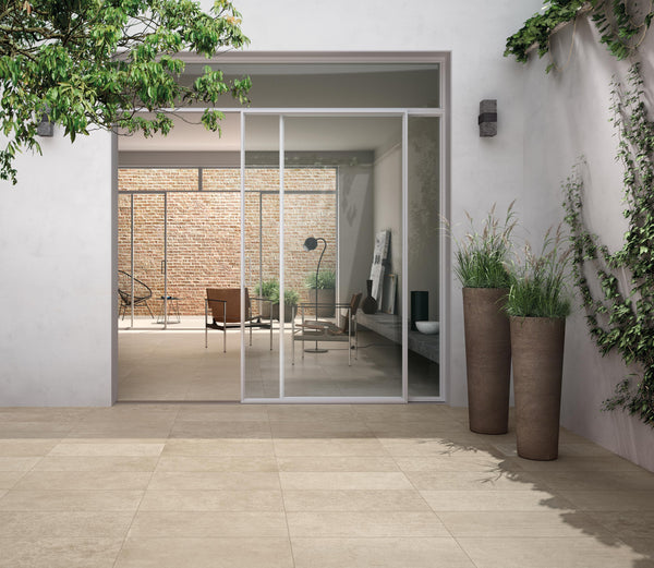 A modern patio with large beige tiles, sliding glass doors, tall planters with green plants, and a view into a stylish indoor living area with natural light and minimalistic decor.