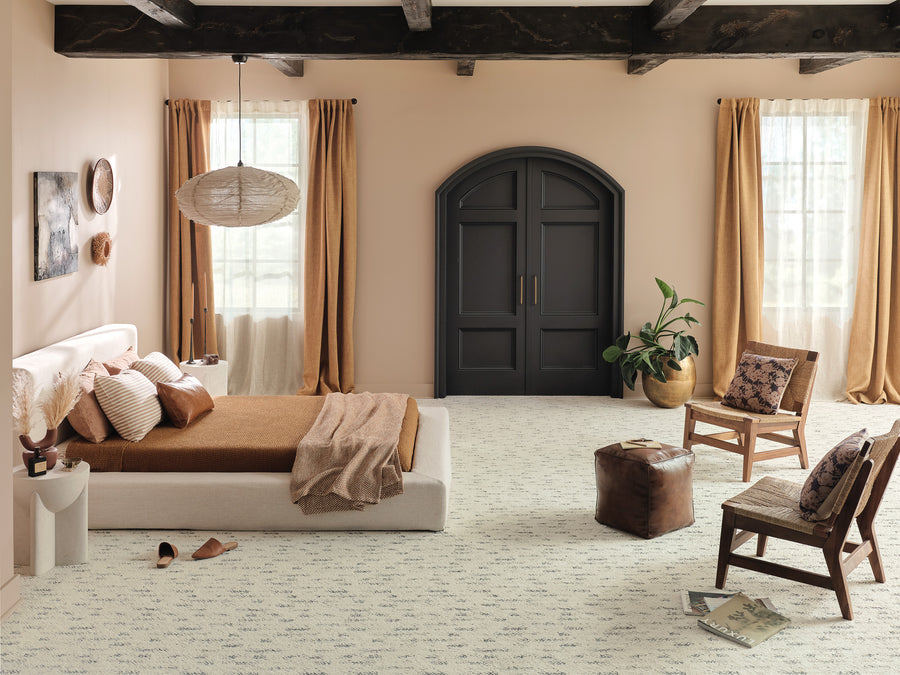 A cozy bedroom with a low bed, brown bedding, and pillows. There are two chairs, a small table, a potted plant, and a brown pouf. Large windows with tan curtains and dark double doors complete the room’s neutral, modern décor.