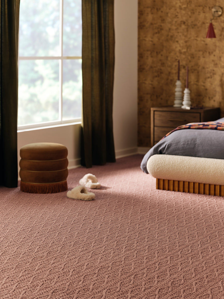 A cozy bedroom with pink textured carpet, a modern cream bed, fluffy slippers on the floor, a brown round ottoman, dark curtains, and a wooden nightstand with decorative objects near a window.