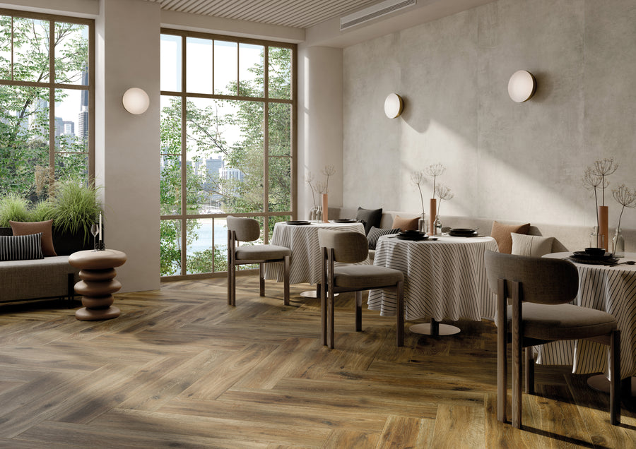 A modern restaurant interior with large windows, wooden herringbone floors, round tables with striped tablecloths, cushioned chairs, minimalist wall lights, and greenery outside the windows.