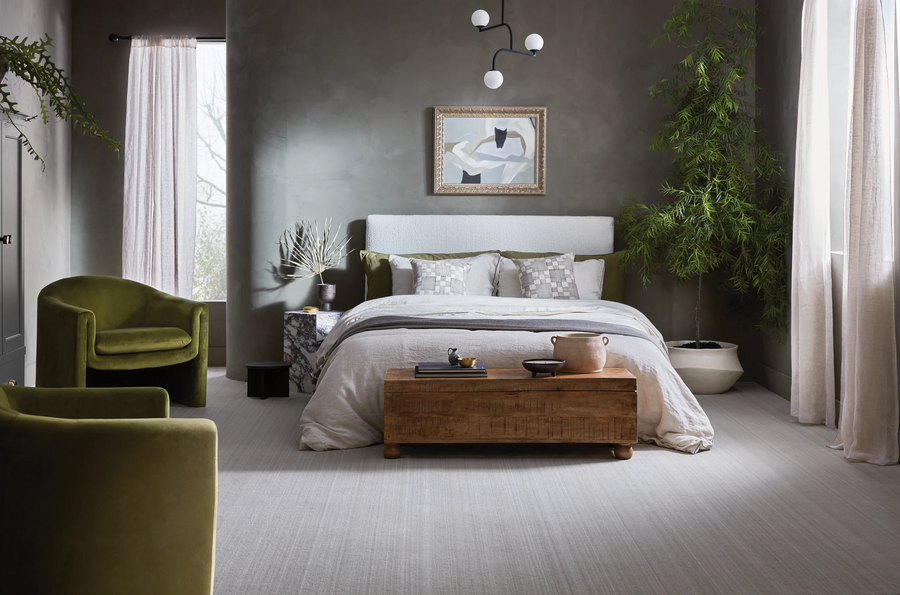 Modern bedroom with a large bed, wooden chest at the foot, green velvet chairs, abstract wall art, potted tree, and soft natural light coming through tall curtains. The decor is neutral with earthy and green accents.