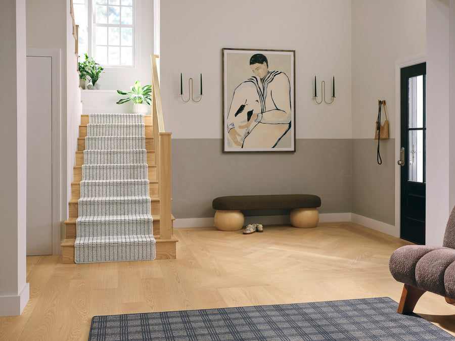 A modern entryway features light wood floors, a staircase with a striped runner, a dark bench, wall art of a person in muted colors, wall sconces, a black door, and houseplants near a window.