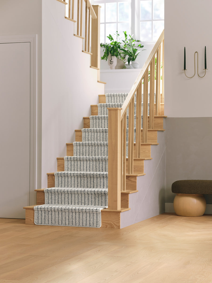 A wooden staircase with a patterned gray and white runner leads up to a bright landing with large windows, potted plants, and natural light. Modern decor includes a wall sconce and a small bench nearby.
