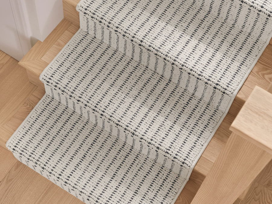 A close-up view of a staircase with light wood steps, covered by a white and black striped woven carpet runner. The pattern features evenly spaced vertical black lines on a white background.