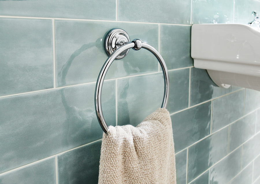 A beige towel hangs on a shiny chrome towel ring mounted on a wall with glossy light blue-green tiles, next to a white sink.