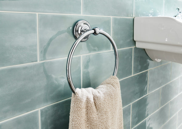 A beige towel hangs on a shiny chrome towel ring mounted on a wall with glossy light blue-green tiles, next to a white sink.