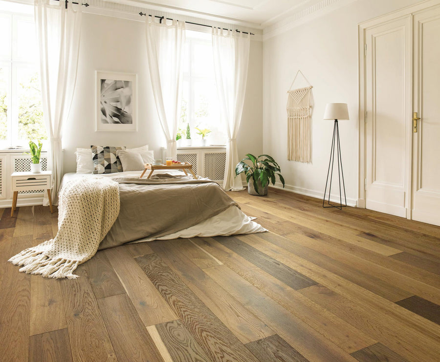 A bright bedroom with large windows, light curtains, a neatly made bed with beige bedding and a knit throw, wooden floors, wall decor, a potted plant, a floor lamp, and a tray on the bed.