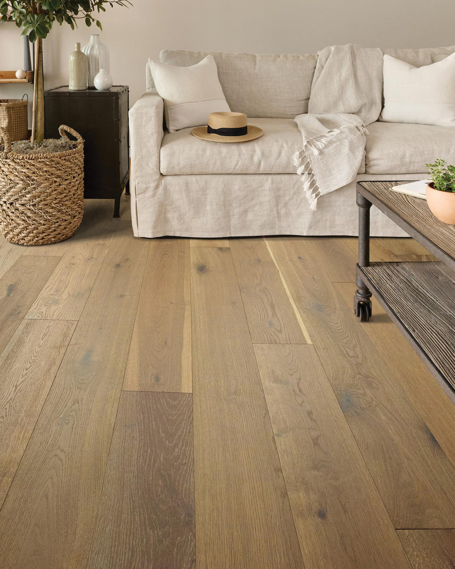 A cozy living room features light wood flooring, a beige sofa with white pillows and a throw blanket, a wicker basket, a black cabinet, and a coffee table with metal legs. A hat rests on the sofa.