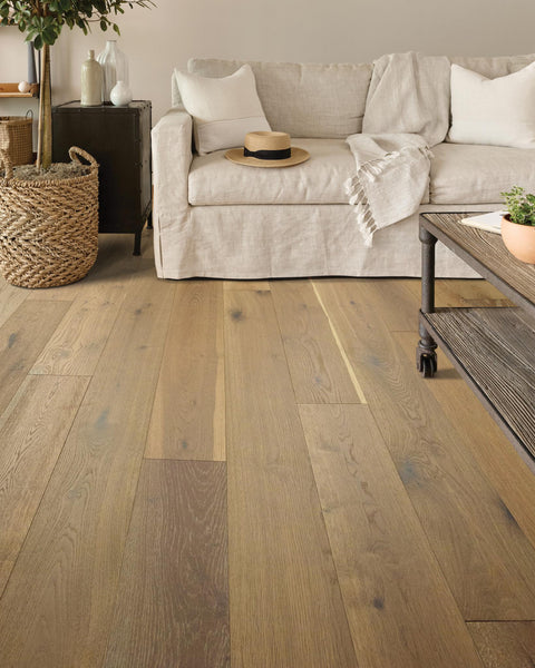 A cozy living room features light wood flooring, a beige sofa with white pillows and a throw blanket, a wicker basket, a black cabinet, and a coffee table with metal legs. A hat rests on the sofa.