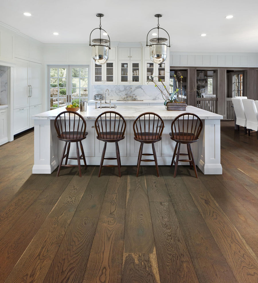Spacious kitchen with white cabinets, a large marble island, four wooden chairs, pendant lights, and wide plank hardwood floors. Dining area with white chairs is visible in the background.