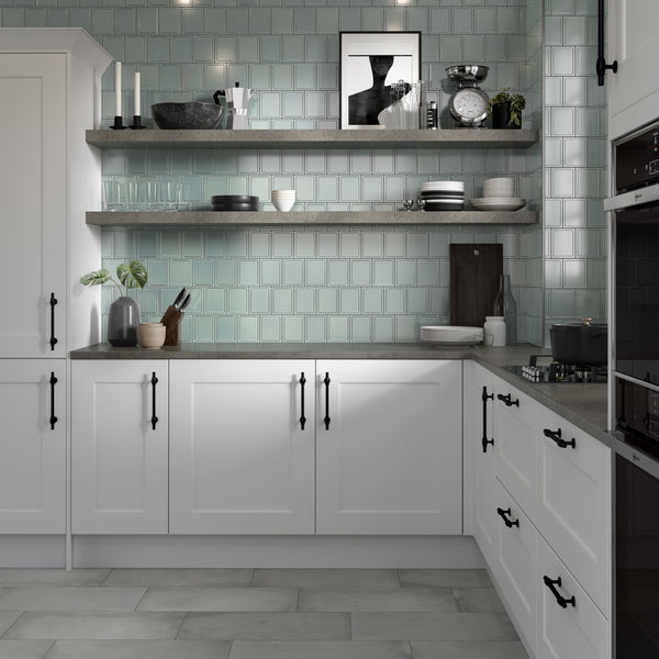 Modern kitchen with white cabinets, black handles, gray countertops, and light blue subway tile backsplash. Open shelves display dishes, bowls, plants, candles, and framed art. Built-in oven and stove are visible.