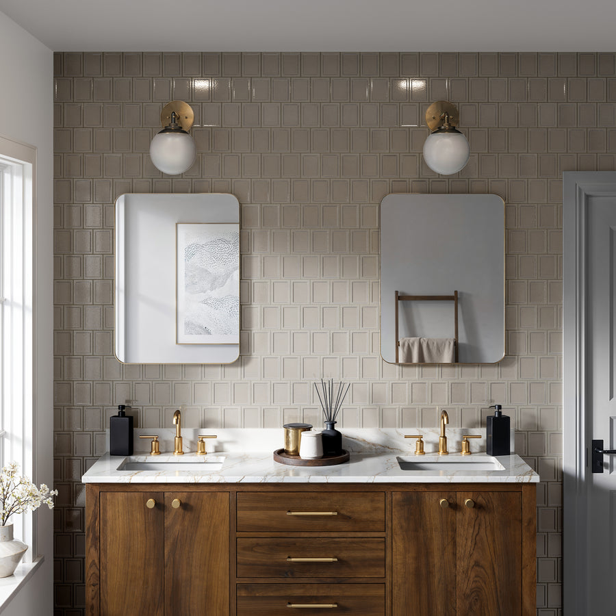 A modern bathroom with a double vanity, marble countertop, wooden cabinets, gold fixtures, two mirrors, wall sconces, and beige tiled walls. A towel rack and window bring in natural light.