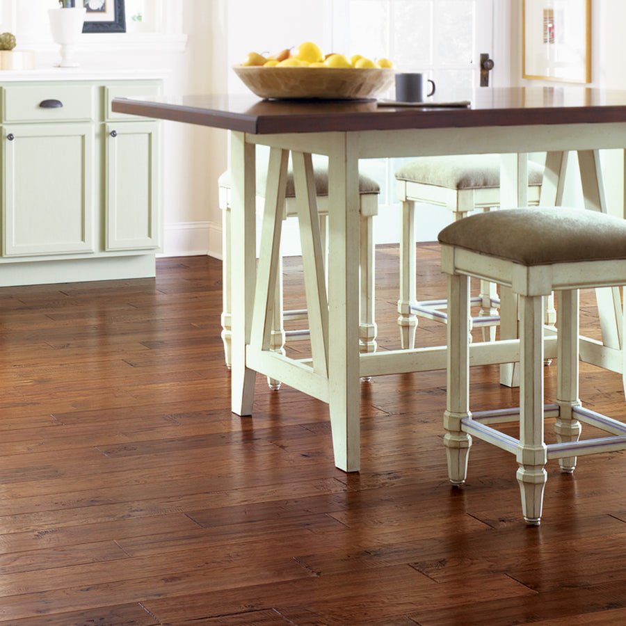 A bright kitchen featuring Deepwood Trail 5″ Hickory Oak Hardwood flooring, a white island table with cushioned stools, and a bowl of lemons atop the countertop. Sunlight streams through a window in the background.