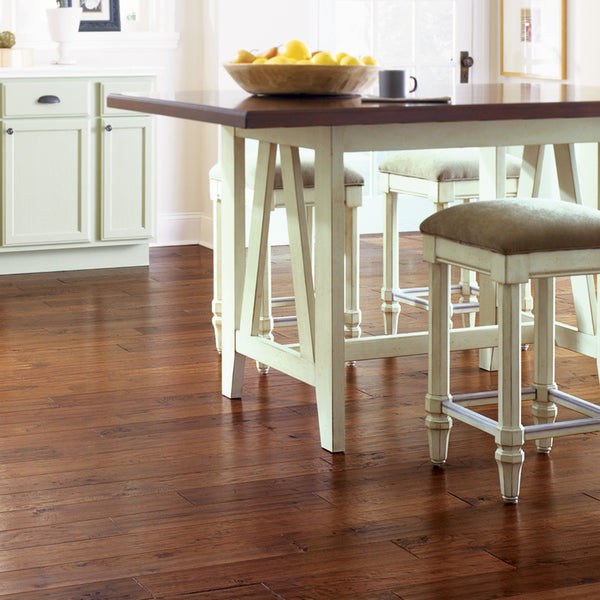 A bright kitchen featuring Deepwood Trail 5″ Hickory Oak Hardwood flooring, a white island table with cushioned stools, and a bowl of lemons atop the countertop. Sunlight streams through a window in the background.