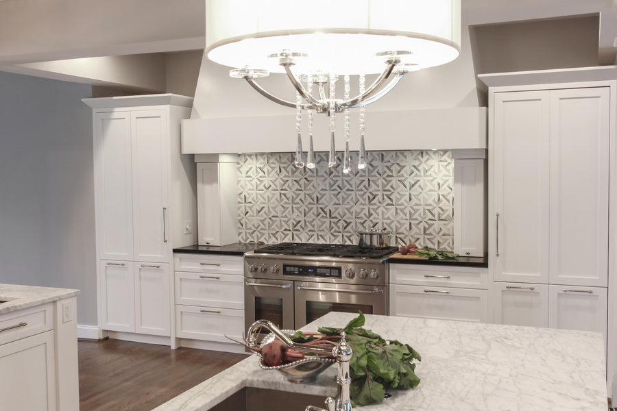 Modern kitchen with white cabinets, marble countertops, stainless steel stove, geometric tile backsplash, and a large chandelier. Fresh vegetables are on the counter near the sink.