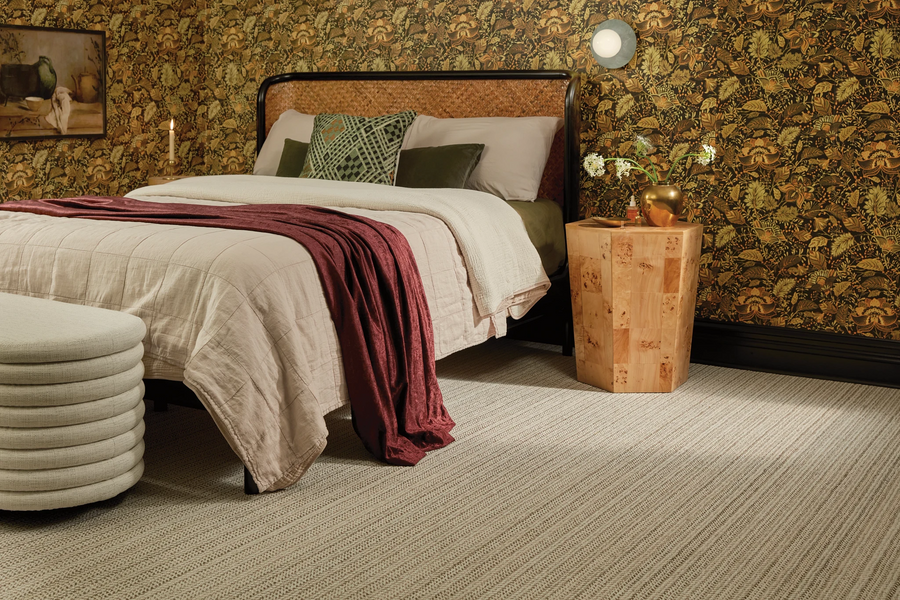 A cozy bedroom with a bed topped with green and beige pillows, a burgundy throw, patterned wallpaper, a wooden nightstand with a vase, a small round wall light, and beige carpet flooring.