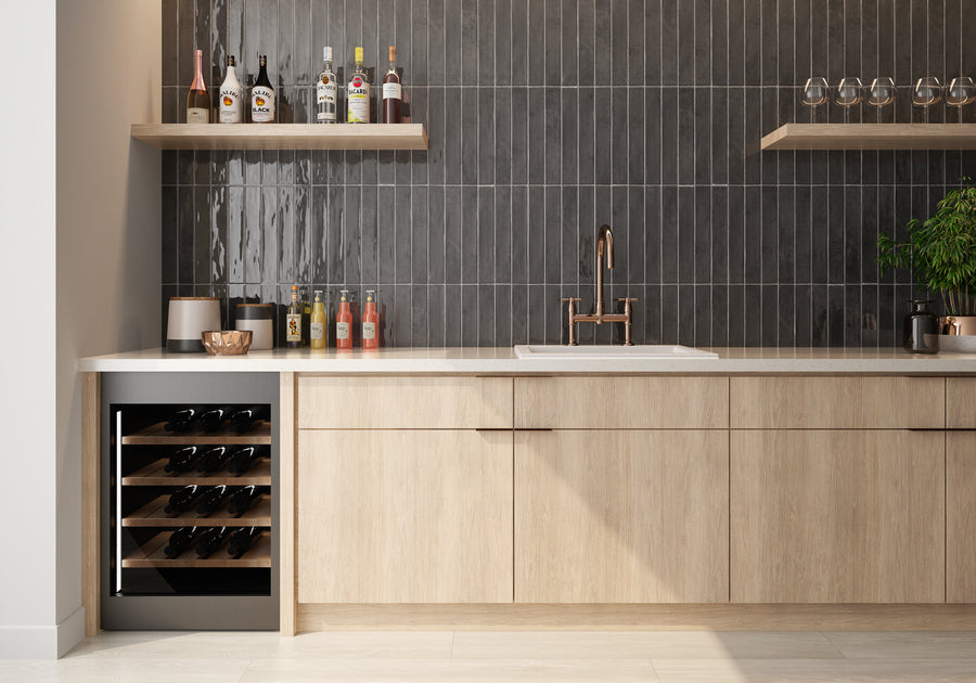 Modern kitchen with light wood cabinets, a built-in wine cooler, open shelves with bottles and glasses, a white sink with a bronze faucet, dark tiled backsplash, and a green plant on the counter.