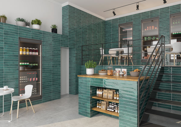 A modern café with green tile walls, shelves of jars and products, a wooden counter with snacks, indoor plants, and a black metal staircase leading to an upper seating area with white chairs and tables.