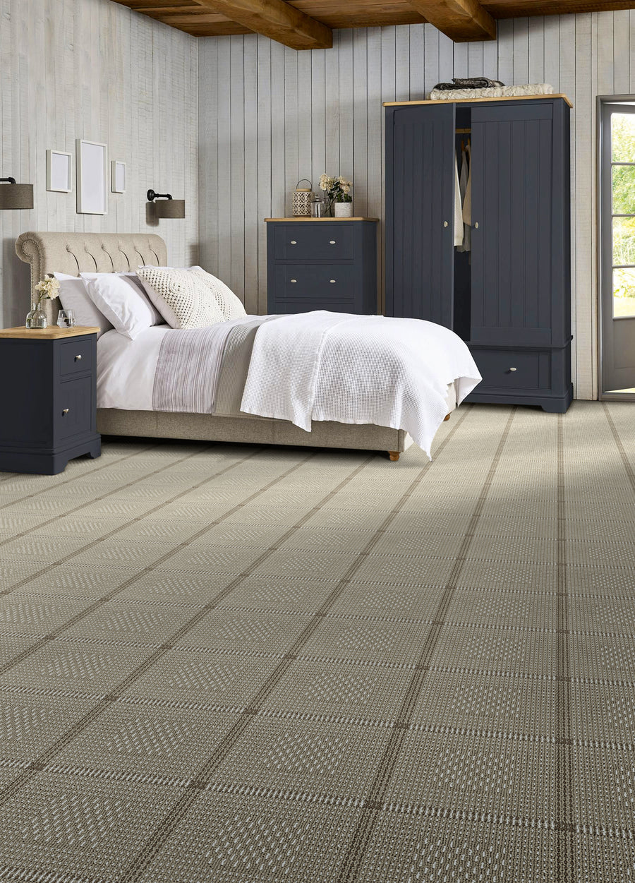 A cozy bedroom with a beige upholstered bed, white bedding, two dark navy nightstands, a matching wardrobe, dresser, and light wood paneled walls. The floor has a light patterned carpet and a wooden ceiling with exposed beams.