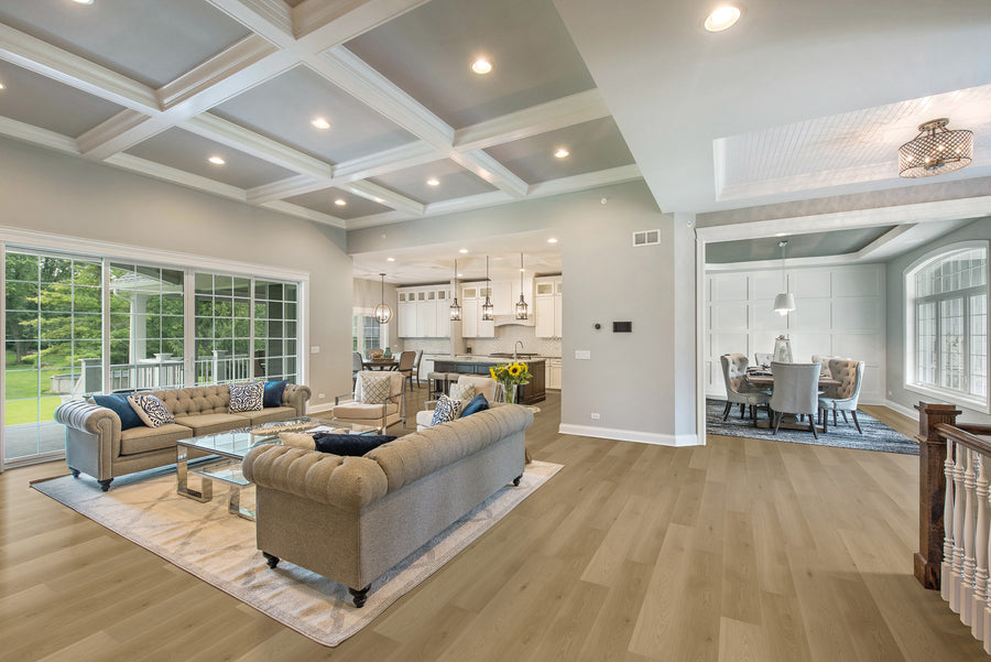 A spacious, modern open-concept living room and kitchen with coffered ceilings, neutral-colored sofas, glass coffee table, wooden floors, large windows, and a dining area in the background.