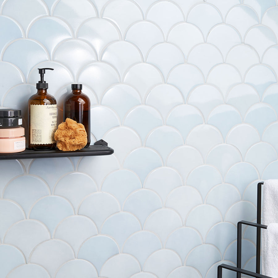A bathroom wall with light blue scallop-patterned tiles. A black shelf holds three bottles, a sponge, and a jar. Part of a black towel rack with a white towel is visible on the right side.