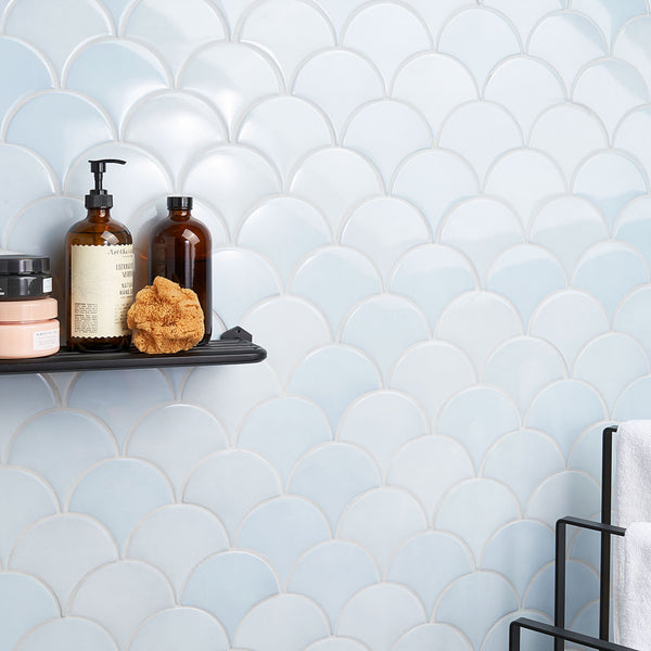 A bathroom wall with light blue scallop-patterned tiles. A black shelf holds three bottles, a sponge, and a jar. Part of a black towel rack with a white towel is visible on the right side.