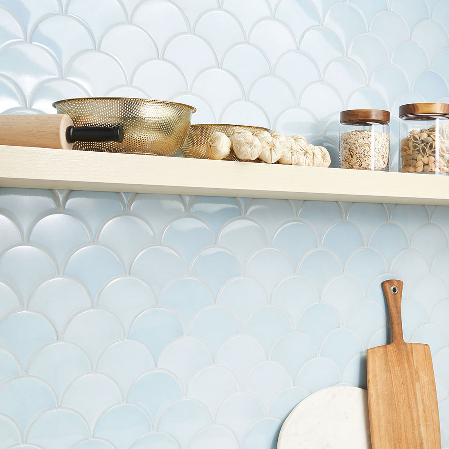 A kitchen shelf holds a gold colander, a rolling pin, bulbs of garlic, and two glass jars with wooden lids in front of a light blue, scallop-patterned tile backsplash. A cutting board leans against the wall below.