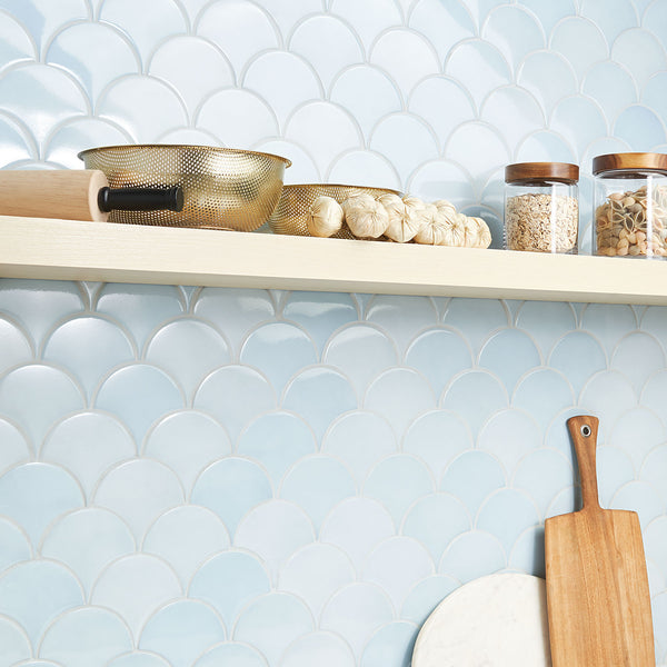 A kitchen shelf holds a gold colander, a rolling pin, bulbs of garlic, and two glass jars with wooden lids in front of a light blue, scallop-patterned tile backsplash. A cutting board leans against the wall below.